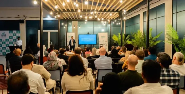 An audience listens to a speaker during an event in a modern, well-lit venue with greenery and branded banners.